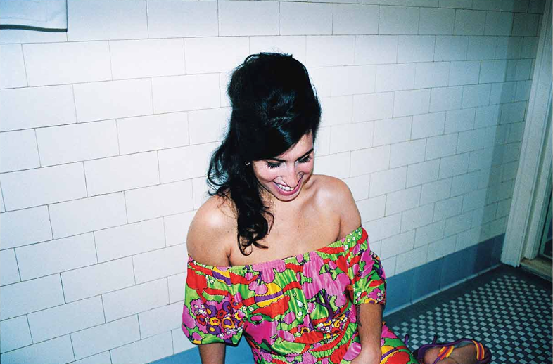A colour film photograph of a woman with black hair styled in a vintage updo, wearing a vibrant off-shoulder floral print top with red, pink, green and orange patterns, seated on a checkered black and white floor against white subway tiles in what…